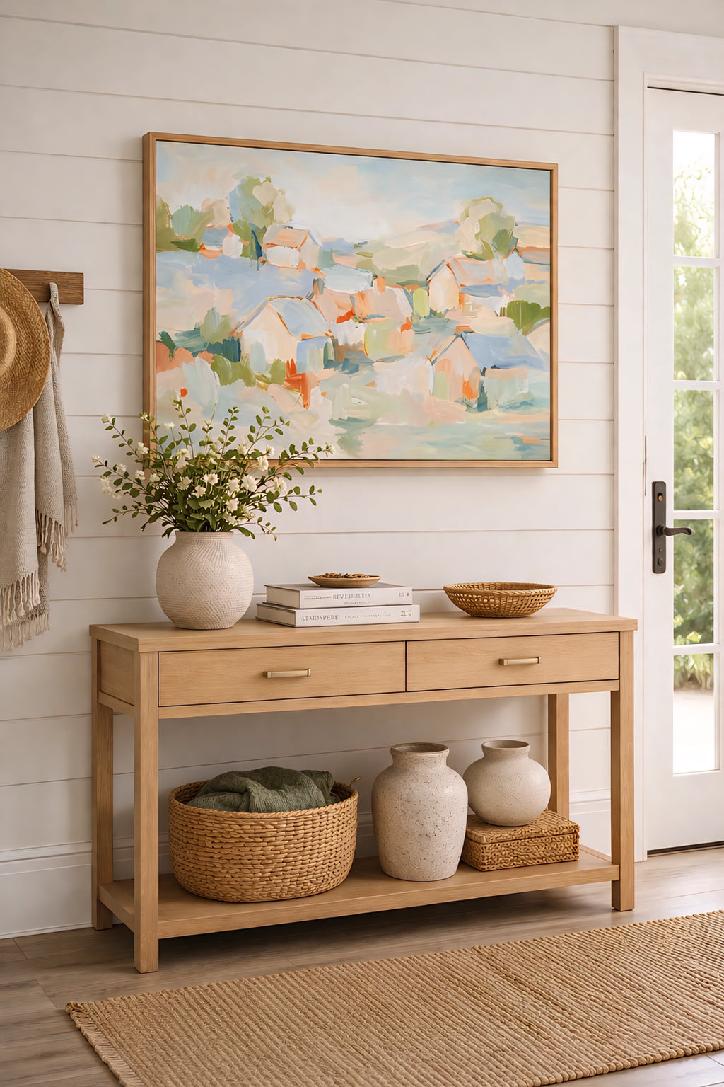Abstract coastal village painting in soft pastel tones displayed in a light wood frame above a console table in a bright entryway with neutral decor, woven textures, and airy modern coastal styling.