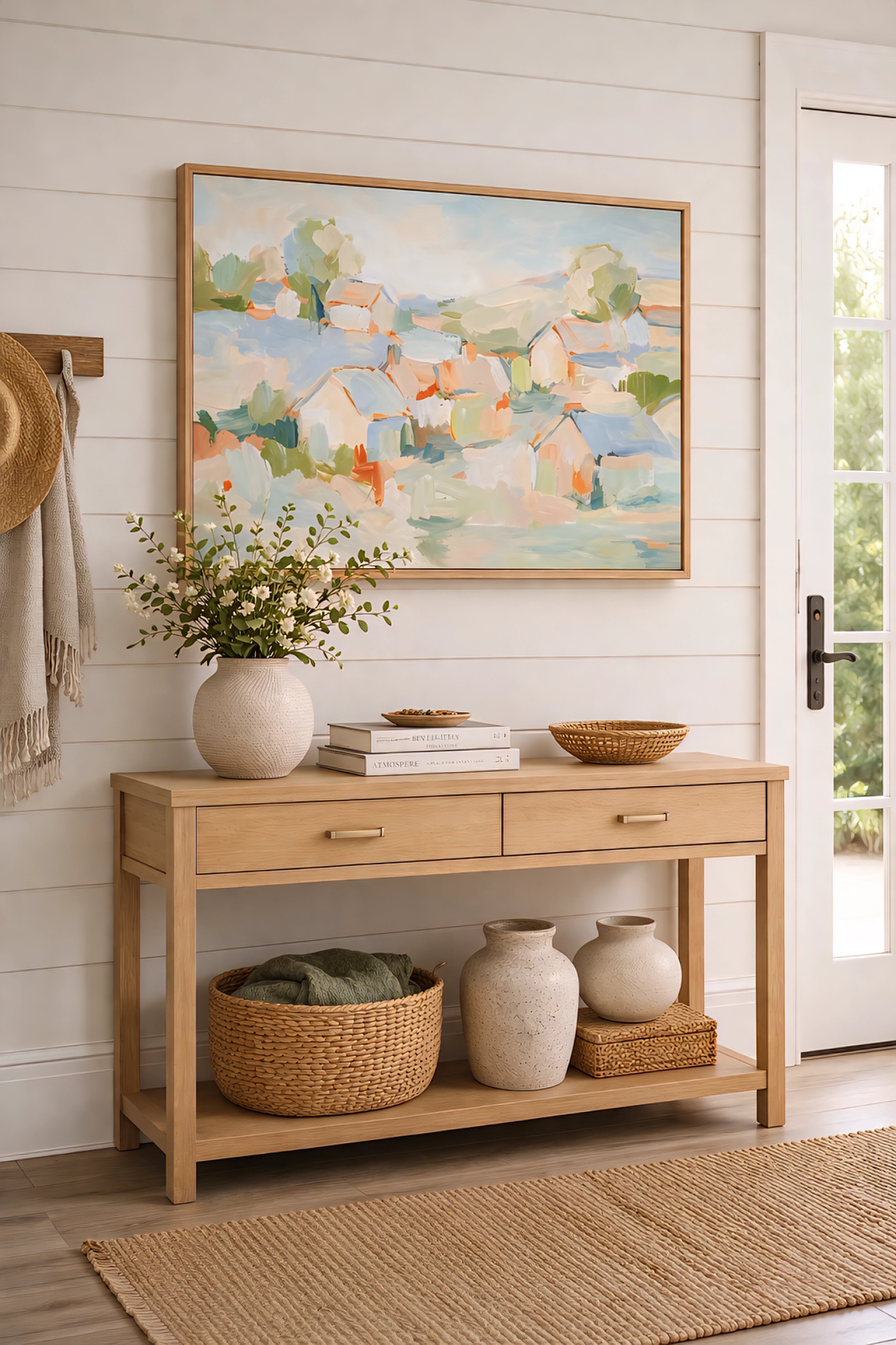 Abstract coastal village painting in soft pastel tones displayed in a light wood frame above a console table in a bright entryway with neutral decor, woven textures, and airy modern coastal styling.