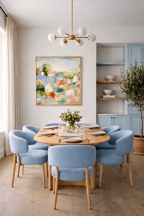 Abstract coastal village painting in soft pastel tones displayed in a light wood frame above a dining table, styled in a bright modern dining room with blue upholstered chairs, warm wood accents, and airy coastal decor.